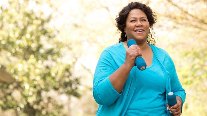 a middle aged Black woman outside, smiling while exercising, walking and carrying hand weights, workout